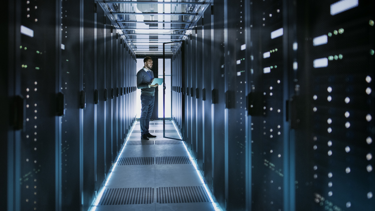 IT Technician Works on Laptop in Colocation Data Centers full of Rack Servers. He Runs Diagnostics and Maintenance, Sets System Up.